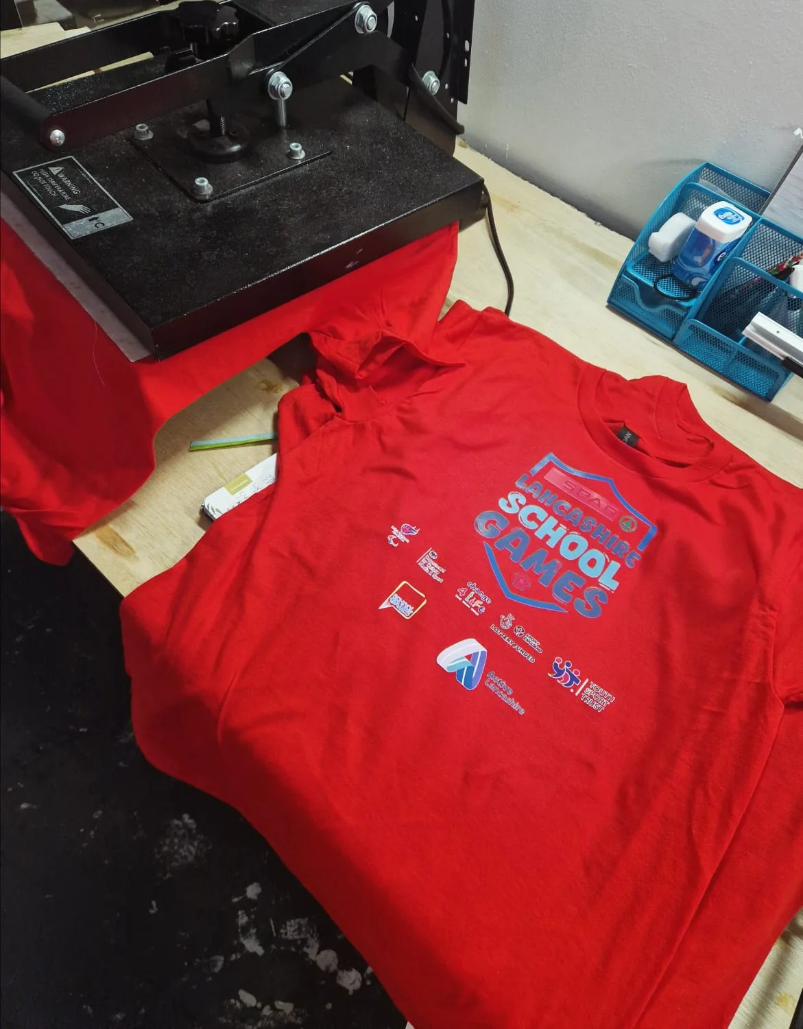 Red school sports top with multiple sponsor logos beside a heat press